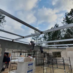 Montaż konstrukcji stalowej dachu na budynku z bloczków betonowych, dwóch spawaczy na rusztowaniu łączy elementy, na pierwszym planie paleta z materiałem Solbet.