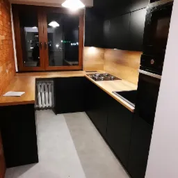 Nowoczesna kuchnia z czarnymi frontami i drewnianym blatem, oświetlona lampą wiszącą, z widocznym fragmentem ściany z czerwonej cegły i grzejnikiem pod oknem.