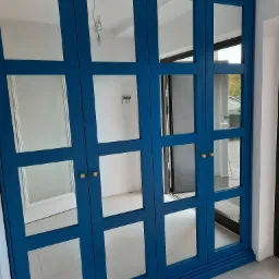 Niebieska szafa na wymiar z lustrzanymi drzwiami i złotymi gałkami, widok na wnętrze pomieszczenia z niedokończonym oświetleniem.