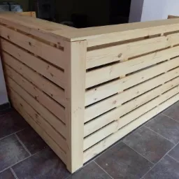 Drewniana lada barowa z jasnego drewna, wykonana z poziomych desek z przerwami, stojąca na ciemnej, ceramicznej podłodze.