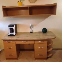 Biurko z drewna w jasnym odcieniu, z szufladami i półkami po prawej stronie, nad nim wisząca półka, na blacie ekspres do kawy, świecznik i zielona kula dekoracyjna.