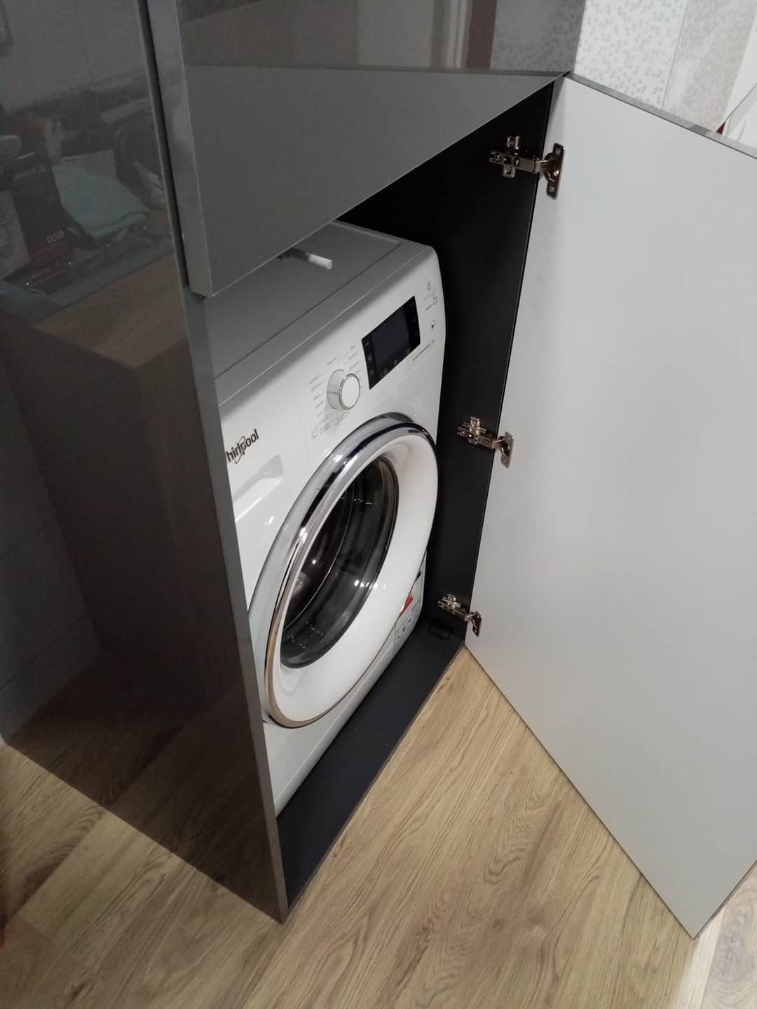 Pralka ukryta w szafce z połyskującymi, grafitowymi frontami i białymi drzwiczkami, widoczne zawiasy.