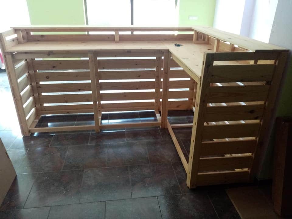 Surowa drewniana konstrukcja baru z poziomych desek, widoczna na tle podłogi z płytek i fragmentu ściany.