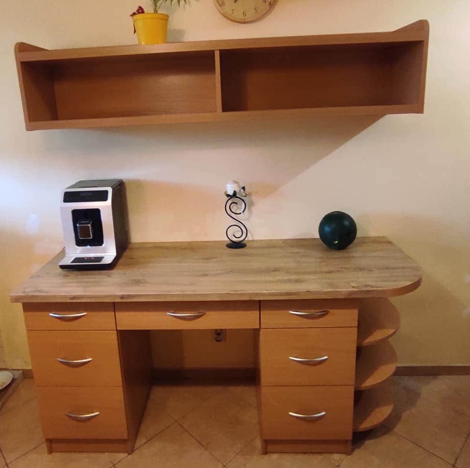 Biurko z drewna w jasnym odcieniu, z szufladami i półkami po prawej stronie, nad nim wisząca półka, na blacie ekspres do kawy, świecznik i zielona kula dekoracyjna.