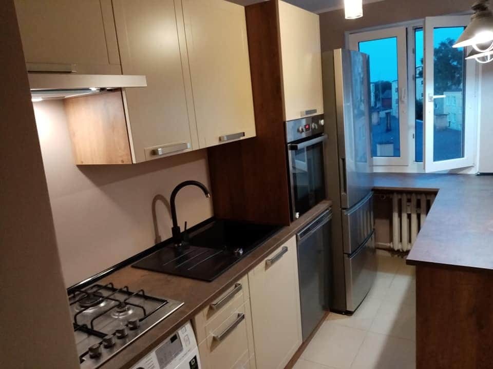 Jasna kuchnia z beżowymi szafkami, czarnym zlewem i kuchenką gazową, widok z boku na okno balkonowe z widokiem na budynki w oddali.