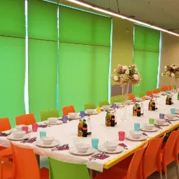 Długi stół bankietowy nakryty białym obrusem, z kolorowymi krzesłami (pomarańczowe i zielone), zastawą stołową, napojami i dekoracjami kwiatowymi w wysokich wazonach, tło stanowią zielone rolety...