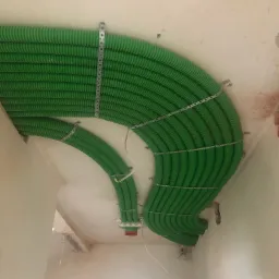 Zielone, karbowane rury wentylacyjne, prawdopodobnie rekuperacyjne, zamocowane do sufitu metalowymi obejmami w surowym pomieszczeniu.