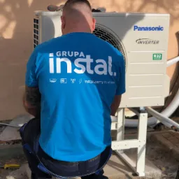 Instalator w niebieskiej koszulce firmy instalacyjnej klęczy przed zewnętrzną jednostką klimatyzacji Panasonic Inverter R32 podczas montażu na betonowym fundamencie.