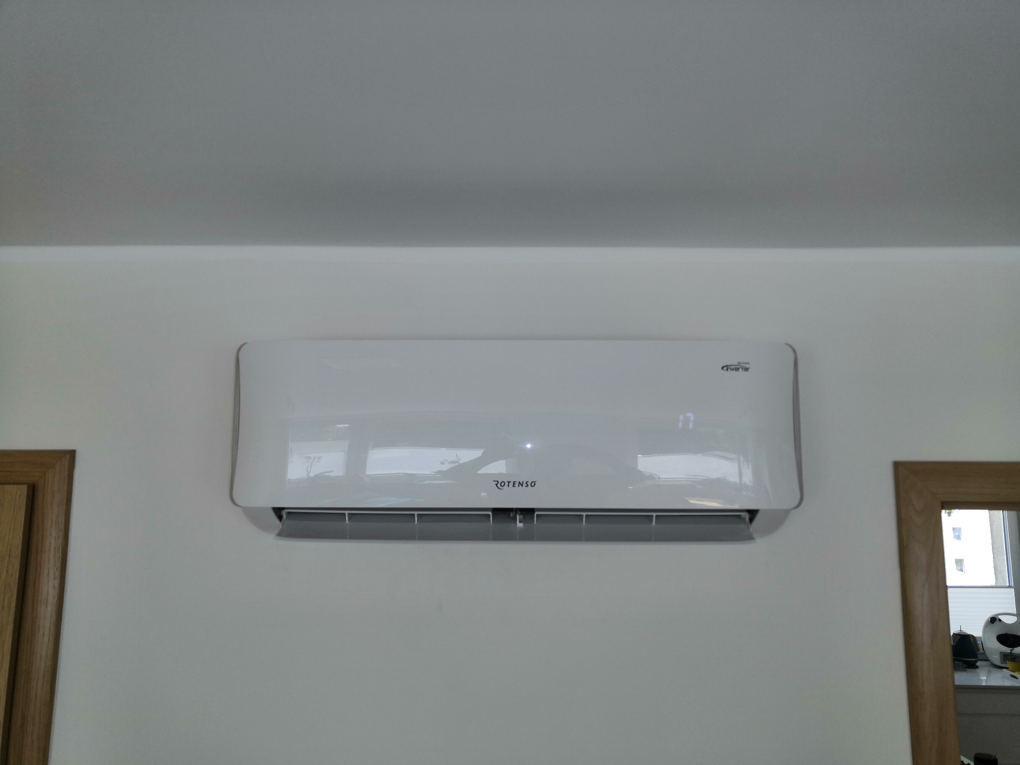 Biały klimatyzator Rotenso Inverter zamontowany na ścianie w pomieszczeniu, widoczne odbicie okna w obudowie, obok fragment ościeżnicy drzwi i lustra.