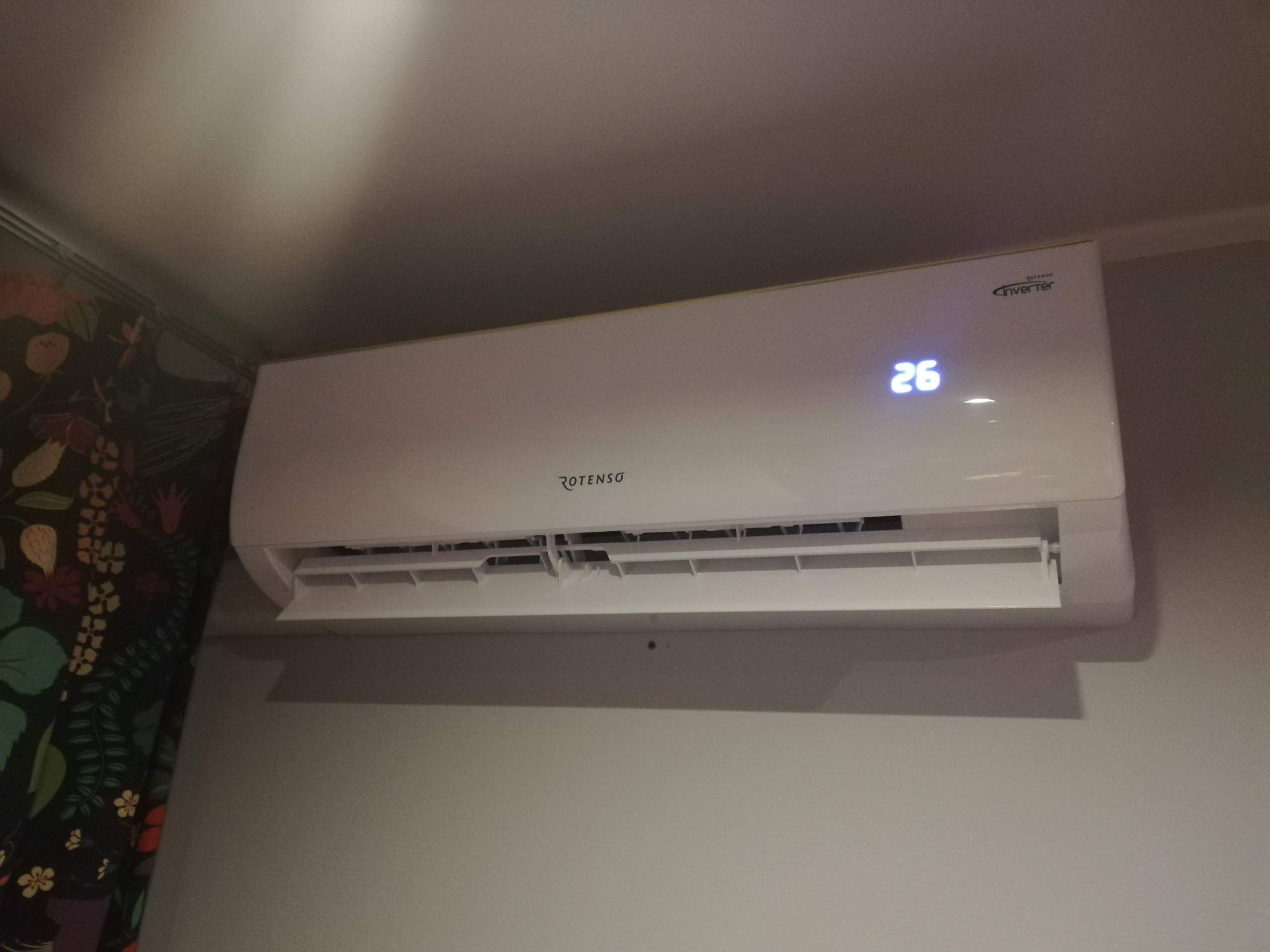 Biała jednostka wewnętrzna klimatyzatora Rotenso z wyświetlaczem temperatury 26 stopni Celsjusza, zamontowana na ścianie obok wzorzystej zasłony.