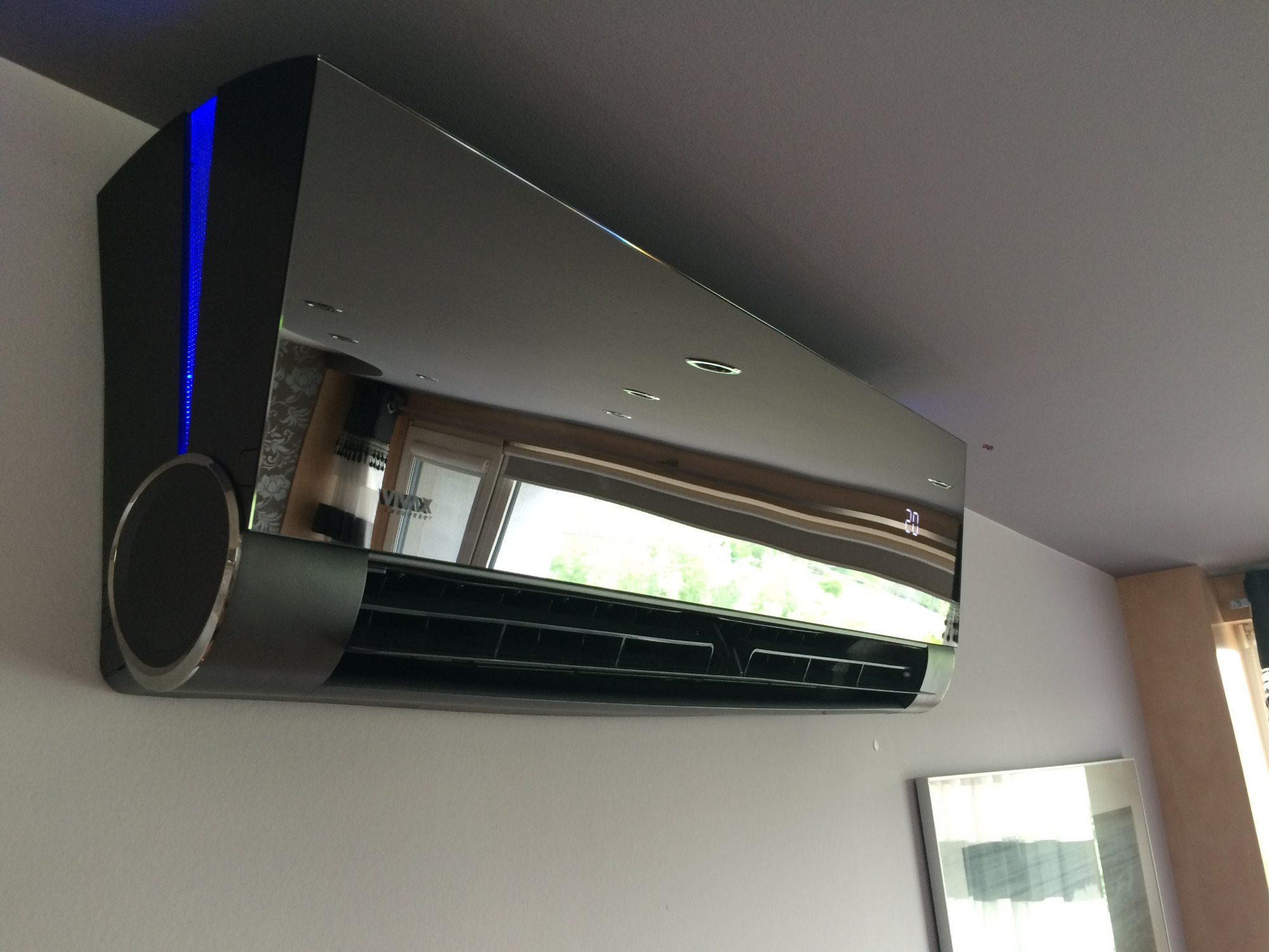 Nowoczesny klimatyzator ścienny z czarną obudową i niebieskim podświetleniem bocznym, zamontowany w jasnym pomieszczeniu z widocznym oknem w odbiciu.