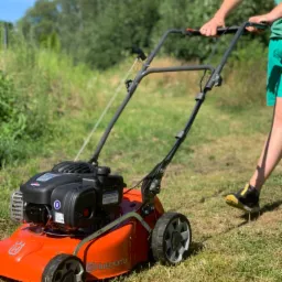Pomarańczowa kosiarka spalinowa Husqvarna prowadzona przez mężczyznę w zielonych spodenkach na tle wysokiej trawy i drzew.