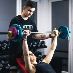 Kobieta ćwiczy wyciskanie sztangi na ławce skośnej pod nadzorem trenera w czarnej koszulce z tatuażem na przedramieniu. Sztanga ma kolorowe obciążenia: niebieskie, różowe i żółte. W tle widoczne są...
