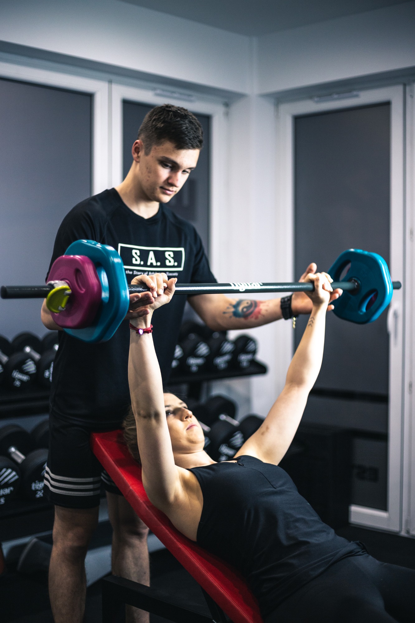 Kobieta ćwiczy wyciskanie sztangi na ławce skośnej pod nadzorem trenera w czarnej koszulce z tatuażem na przedramieniu. Sztanga ma kolorowe obciążenia: niebieskie, różowe i żółte. W tle widoczne są...