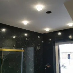 Nowoczesna łazienka z czarnymi płytkami imitującymi marmur, białym blatem z umywalką nablatową i oświetleniem punktowym w suficie, widoczne kable elektryczne wystające ze ściany.