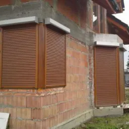 Brązowe rolety zewnętrzne adaptacyjne zamontowane na surowej elewacji z czerwonej cegły dziurawki, widoczne białe prowadnice i fragment dachu domu jednorodzinnego.