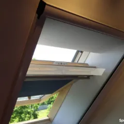Okna dachowe z zamontowanymi roletami i moskitierą, widok na zewnątrz przez otwarte okno.
