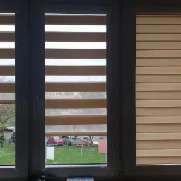 Zamknięte rolety dzień noc w trzech oknach, widok na zewnątrz z zielenią i samochodami, dekoracja z kulkami świetlnymi przy oknie po prawej.