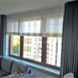 Salon z szarym narożnikiem, oknem z trzema segmentami z roletami rzymskimi w biało-szare pasy oraz szarymi zasłonami po bokach, widok na budynki za oknem.