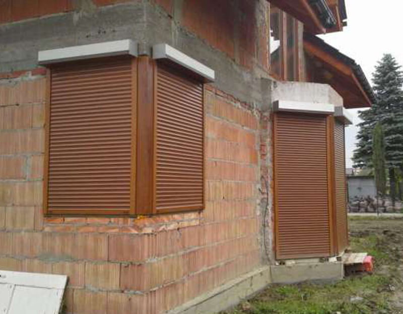 Brązowe rolety zewnętrzne adaptacyjne zamontowane na surowej elewacji z czerwonej cegły dziurawki, widoczne białe prowadnice i fragment dachu domu jednorodzinnego.