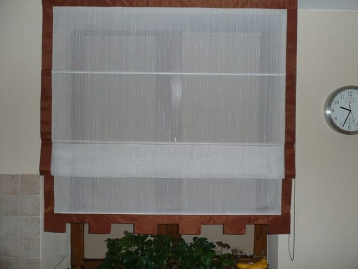 Roleta rzymska z brązowym obszyciem, częściowo opuszczona, widoczny zegar na ścianie i roślina w doniczce w tle.