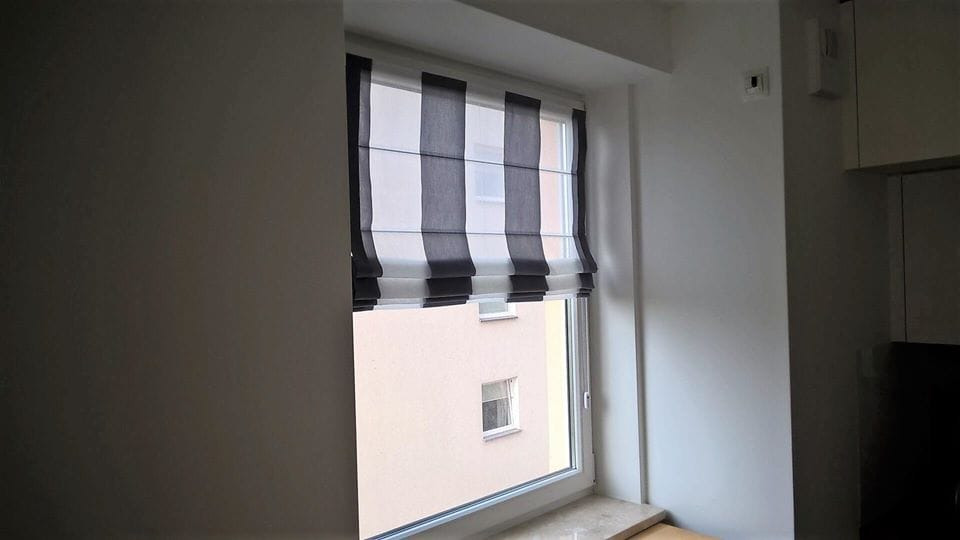 Roleta rzymska w biało-szare pasy, częściowo podniesiona, zamontowana w oknie z widokiem na budynek.