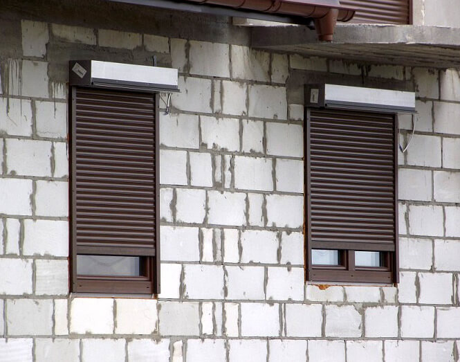 Dwa brązowe okna z roletami zewnętrznymi adaptacyjnymi zamontowane w surowej ścianie z białych bloczków betonowych w trakcie budowy.