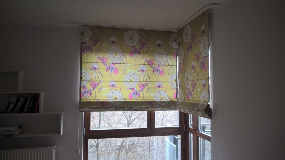 Złożona roleta rzymska w kwiatowy wzór na oknie narożnym, widok z wnętrza pokoju z półkami na książki po lewej stronie.