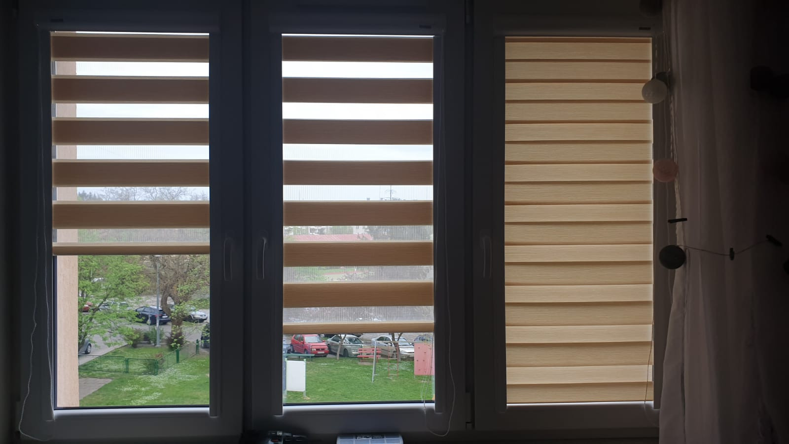 Zamknięte rolety dzień noc w trzech oknach, widok na zewnątrz z zielenią i samochodami, dekoracja z kulkami świetlnymi przy oknie po prawej.