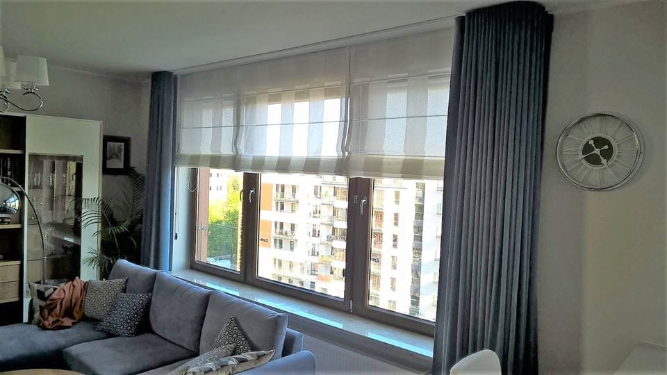 Salon z szarym narożnikiem, oknem z trzema segmentami z roletami rzymskimi w biało-szare pasy oraz szarymi zasłonami po bokach, widok na budynki za oknem.