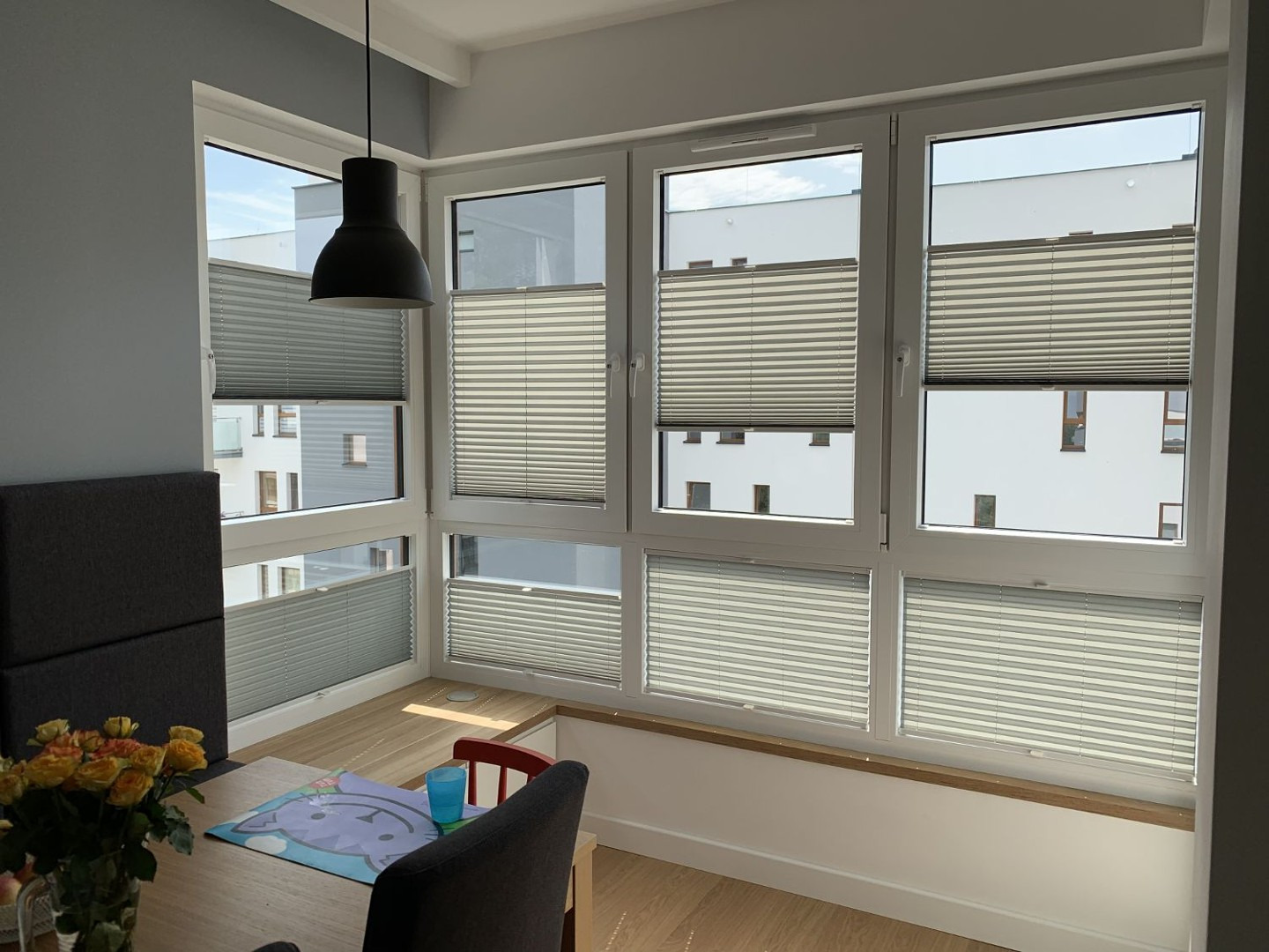 Jasne pomieszczenie z oknami wyposażonymi w szare, poziome osłony okienne; widoczne częściowo odsłonięte okna z widokiem na budynki na zewnątrz.