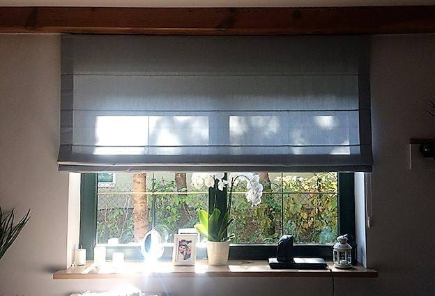 Szara roleta rzymska zamontowana w oknie z widokiem na zieleń ogrodu, na parapecie dekoracje: biała orchidea w doniczce, ramka ze zdjęciem, ozdobny lampion.