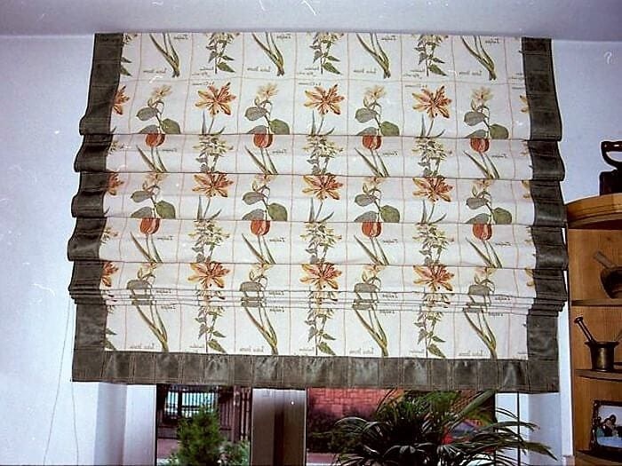 Zasłonięte okno roletą rzymską z motywem kwiatowym, wykończoną ciemnozieloną lamówką, widok od wewnątrz, w tle fragment pokoju z drewnianymi meblami.