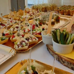 Stół z różnorodnymi przekąskami: sałatki w łódeczkach z ciasta, koreczki, mini-szaszłyki, pieczywo na drewnianej desce, ogórki i dip w białych miseczkach, wszystko na złotych tacach.