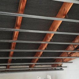 WROT BUD WRÓTNIAK ŁUKASZ Usługi Remontowo Budowlane - Konstrukcja metalowa podwieszanego sufitu zamontowana do drewnianych belek stropowych, widoczne czarne płyty izolacyjne i fragment instalacji elektrycznej.