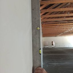 WROT BUD WRÓTNIAK ŁUKASZ Usługi Remontowo Budowlane - Sprawdzanie pionu ściany za pomocą poziomicy w surowym wnętrzu z widoczną drewnianą konstrukcją stropu.