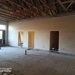 WROT BUD WRÓTNIAK ŁUKASZ Usługi Remontowo Budowlane - Wnętrze w trakcie tynkowania maszynowego, widoczne ściany pokryte świeżym tynkiem, drewniana konstrukcja dachu, surowa posadzka i wystające kable elektryczne.