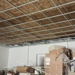 WROT BUD WRÓTNIAK ŁUKASZ Usługi Remontowo Budowlane - Montaż sufitu podwieszanego z widoczną konstrukcją metalową i izolacją akustyczną w przestrzeni biurowej, z zapasem materiałów budowlanych w tle.