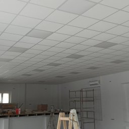 WROT BUD WRÓTNIAK ŁUKASZ Usługi Remontowo Budowlane - Pracownik w kombinezonie malarskim stoi na podwyższeniu w przestronnym pomieszczeniu z kasetonowym sufitem, przygotowując się do prac wykończeniowych. W tle widoczne rusztowania i materiały budowlane.