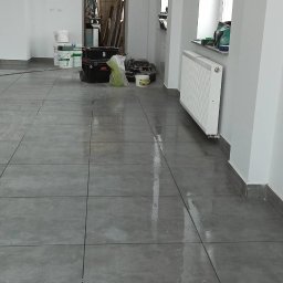 WROT BUD WRÓTNIAK ŁUKASZ Usługi Remontowo Budowlane - Świeżo położona, błyszcząca posadzka z szarych płytek w dużym formacie, z widocznymi śladami wilgoci po czyszczeniu, w tle białe ściany i grzejniki, widoczne narzędzia i materiały budowlane.
