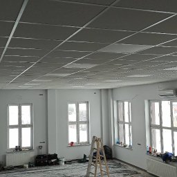 WROT BUD WRÓTNIAK ŁUKASZ Usługi Remontowo Budowlane - Sufit podwieszany kasetonowy z widoczną konstrukcją metalową w jasnym pomieszczeniu biurowym z kilkoma oknami, zabezpieczonym folią malarską na podłodze i drabiną.