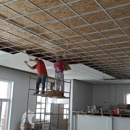 WROT BUD WRÓTNIAK ŁUKASZ Usługi Remontowo Budowlane - Montaż sufitu podwieszanego kasetonowego w pomieszczeniu biurowym. Dwóch pracowników na rusztowaniu instaluje białe płyty w metalowej konstrukcji. Część sufitu już zamontowana, widoczna instalacja...