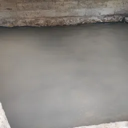 Świeżo wylana, gładka, szara posadzka w pomieszczeniu z surowymi, ceglanymi ścianami, zabezpieczonymi czarną folią przy styku z podłogą.