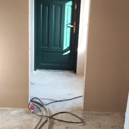 WROT BUD WRÓTNIAK ŁUKASZ Usługi Remontowo Budowlane - Wnętrze w trakcie remontu: świeżo otynkowane ściany w beżowym odcieniu, widoczny fragment zielonych drzwi z ościeżnicą oraz przewody elektryczne na betonowej posadzce.