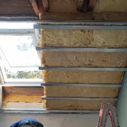 Montaż izolacji z wełny mineralnej w metalowym stelażu sufitu podwieszanego, widoczne okno dachowe i fragment drabiny