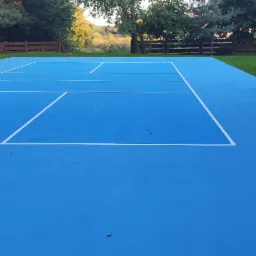 Malowanie nawierzchni boiska do tenisa na kolor niebieski, widoczni dwaj mężczyźni podczas montażu siatki, białe linie wyznaczające pole gry.