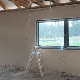 WROT BUD WRÓTNIAK ŁUKASZ Usługi Remontowo Budowlane - Wnętrze w trakcie prac wykończeniowych: ściany po nałożeniu gładzi, widoczne puszki elektryczne, drabina aluminiowa, częściowo odsłonięta konstrukcja drewniana dachu, okno z widokiem na zieleń.