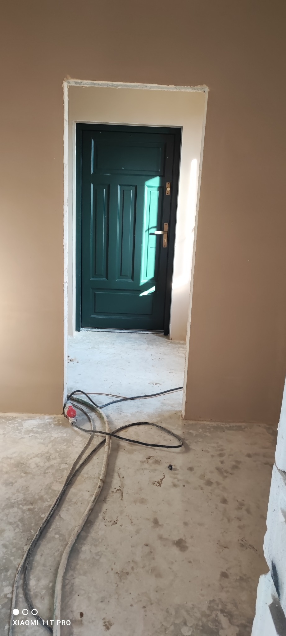 Wnętrze w trakcie remontu: świeżo otynkowane ściany w beżowym odcieniu, widoczny fragment zielonych drzwi z ościeżnicą oraz przewody elektryczne na betonowej posadzce.