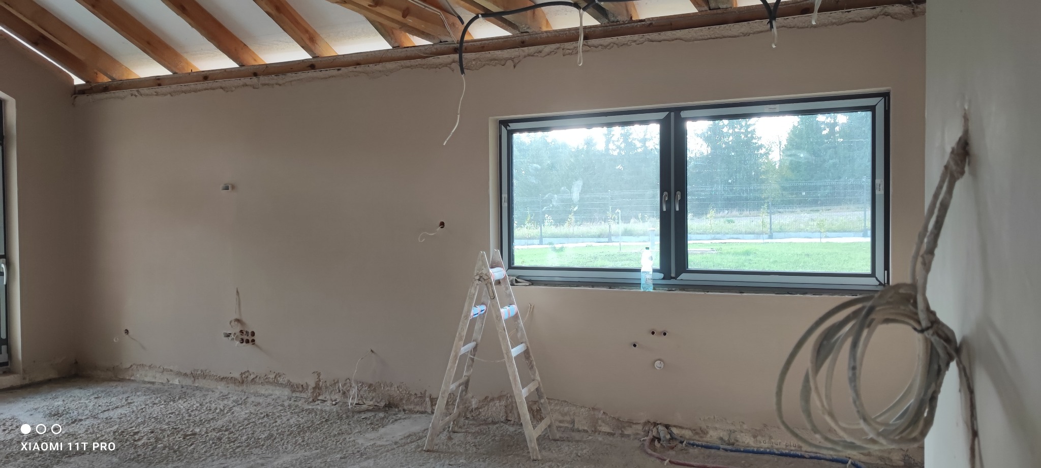 Wnętrze w trakcie prac wykończeniowych: ściany po nałożeniu gładzi, widoczne puszki elektryczne, drabina aluminiowa, częściowo odsłonięta konstrukcja drewniana dachu, okno z widokiem na zieleń.