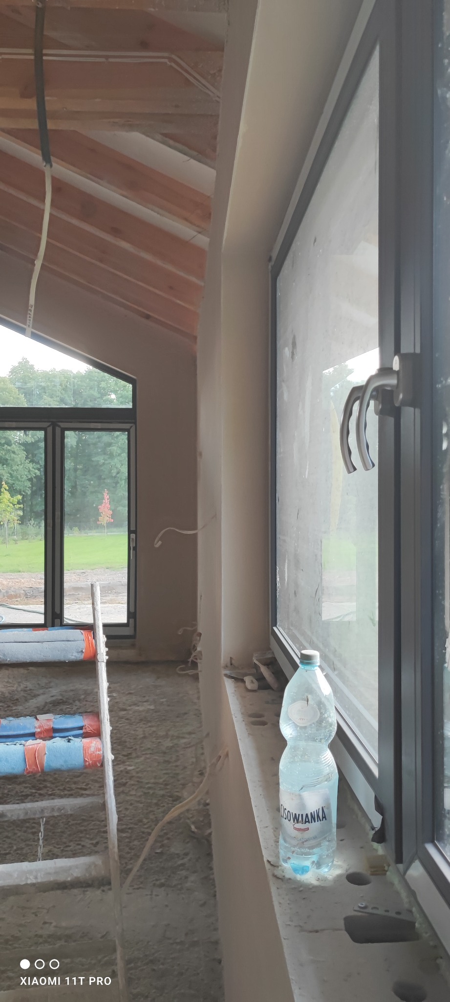 Wnętrze w trakcie prac wykończeniowych: ściany po nałożeniu tynku, widoczne okno z czarną ramą, drabina oparta o ścianę i butelka wody mineralnej na parapecie.